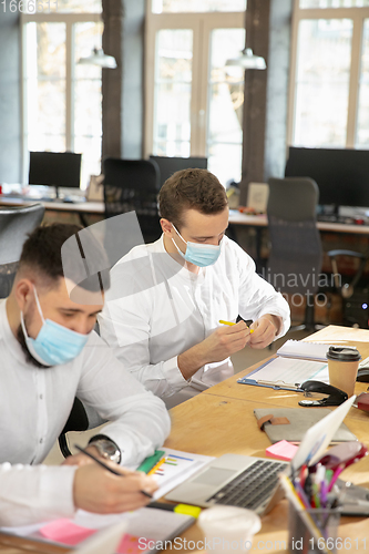 Image of Young caucasian colleagues working together in a office using modern devices and gadgets during quarantine. Wearing protective face masks