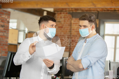 Image of Young caucasian colleagues working together in a office using modern devices and gadgets during quarantine. Wearing protective face masks