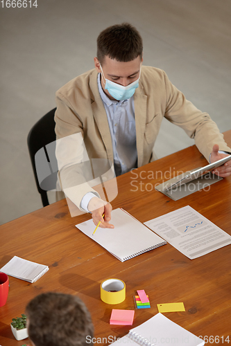 Image of High angle view colleagues working together in face masks during quarantine in a office using modern devices and gadgets during creative meeting