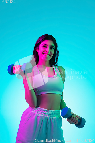 Image of Brunette beautiful woman\'s portrait isolated on blue studio background in multicolored neon light. Training with dumbbells