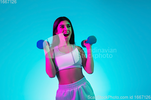 Image of Brunette beautiful woman\'s portrait isolated on blue studio background in multicolored neon light. Training with dumbbells