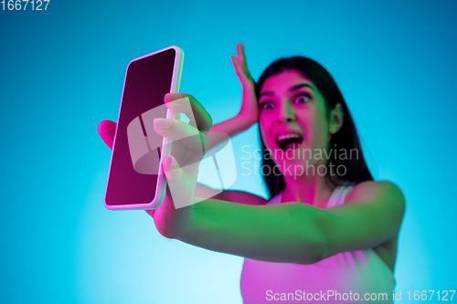 Image of Brunette beautiful woman\'s portrait isolated on blue studio background in multicolored neon light. Showing blank phone\'s screen