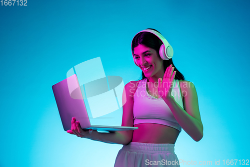 Image of Brunette beautiful woman\'s portrait isolated on blue studio background in multicolored neon light. Model with headphones and laptop