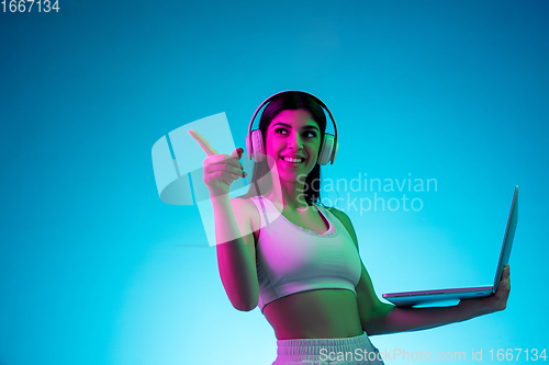 Image of Brunette beautiful woman\'s portrait isolated on blue studio background in multicolored neon light. Model with headphones and laptop