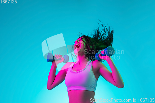 Image of Brunette beautiful woman\'s portrait isolated on blue studio background in multicolored neon light. Training with dumbbells