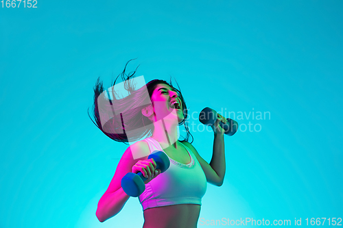 Image of Brunette beautiful woman\'s portrait isolated on blue studio background in multicolored neon light. Training with dumbbells