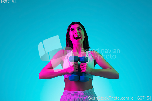 Image of Brunette beautiful woman\'s portrait isolated on blue studio background in multicolored neon light. Training with dumbbells