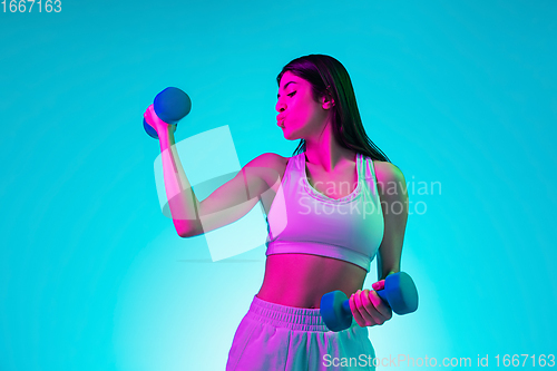Image of Brunette beautiful woman\'s portrait isolated on blue studio background in multicolored neon light. Training with dumbbells
