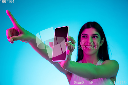 Image of Brunette beautiful woman\'s portrait isolated on blue studio background in multicolored neon light. Showing blank phone\'s screen