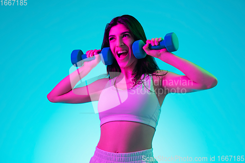 Image of Brunette beautiful woman\'s portrait isolated on blue studio background in multicolored neon light. Training with dumbbells