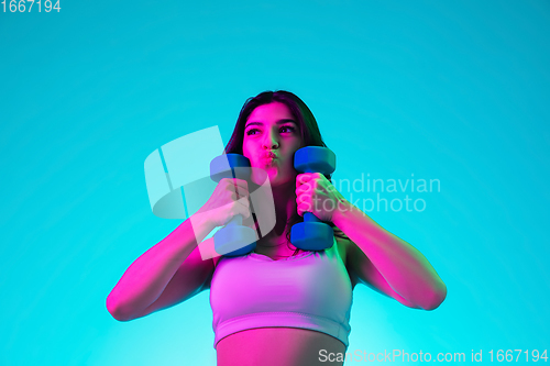 Image of Brunette beautiful woman\'s portrait isolated on blue studio background in multicolored neon light. Training with dumbbells