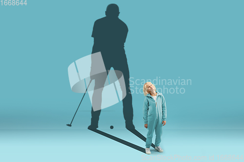 Image of Childhood and dream about big and famous future. Conceptual image with boy and shadow of fit male golf player on blue background