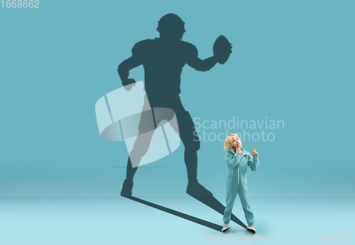 Image of Childhood and dream about big and famous future. Conceptual image with boy and shadow of fit male american football player on blue background