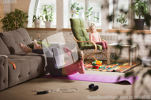 Image of Young woman exercising fitness, aerobic, yoga at home, sporty lifestyle. Getting active while her child playing on the background, home gym.
