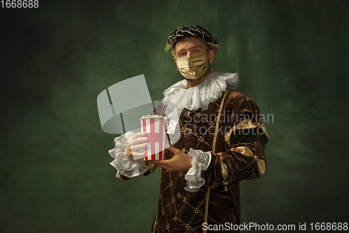 Image of Portrait of medieval young man in vintage clothing and golden face mask standing on dark background.