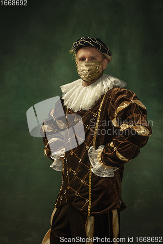 Image of Portrait of medieval young man in vintage clothing and golden face mask standing on dark background.