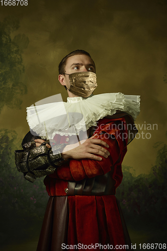 Image of Portrait of medieval young man in vintage clothing and golden face mask standing on dark background.