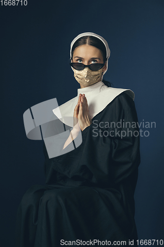 Image of Medieval young woman as a nun in vintage clothing and golden face mask on dark blue background. Concept of comparison of eras, protection from covid