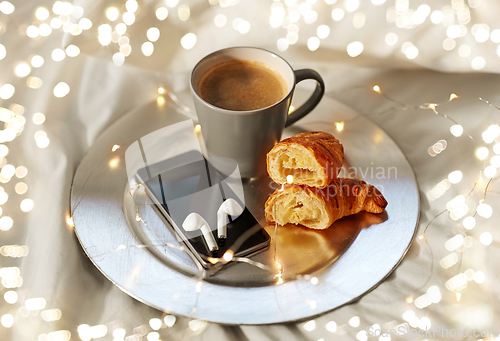 Image of smartphone, earphones, coffee and croissant in bed