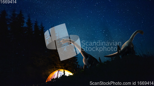 Image of Dinosaurs huge higth walking through the night forest comes across tourists group in travel tent