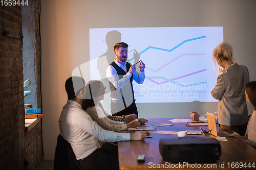 Image of Male speaker giving presentation in hall at workshop. Audience or conference hall. Participants, co-workers listening at the table.