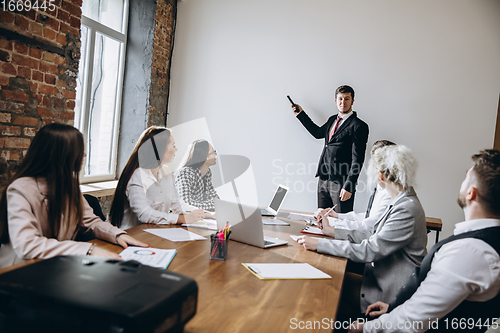 Image of Male speaker giving presentation in hall at workshop. Audience or conference hall. Participants, co-workers listening at the table.