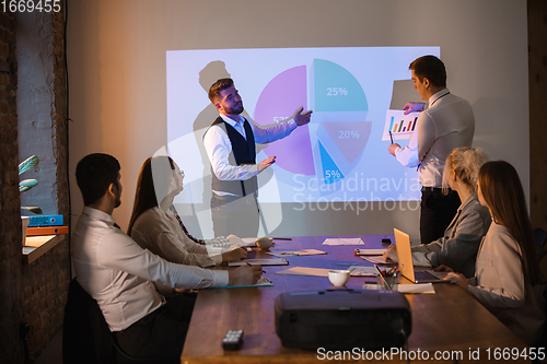 Image of Male speaker giving presentation in hall at workshop. Audience or conference hall. Participants, co-workers listening at the table.