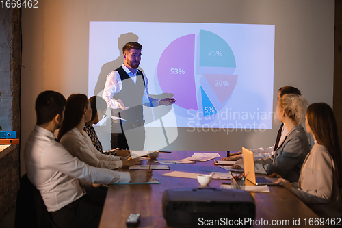 Image of Male speaker giving presentation in hall at workshop. Audience or conference hall. Participants, co-workers listening at the table.