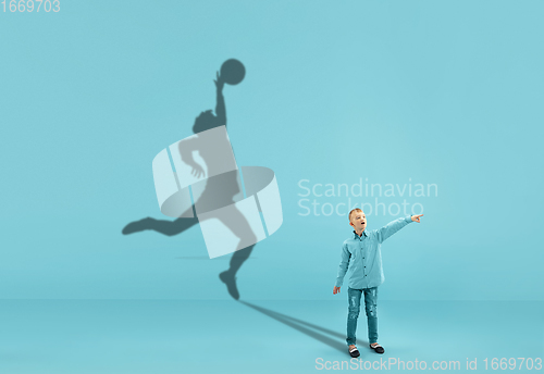 Image of Childhood and dream about big and famous future. Conceptual image with boy and shadow of fit male basketball player on blue background