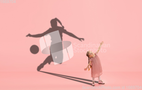 Image of Childhood and dream about big and famous future. Conceptual image with girl and shadow of fit female football player on coral pink background