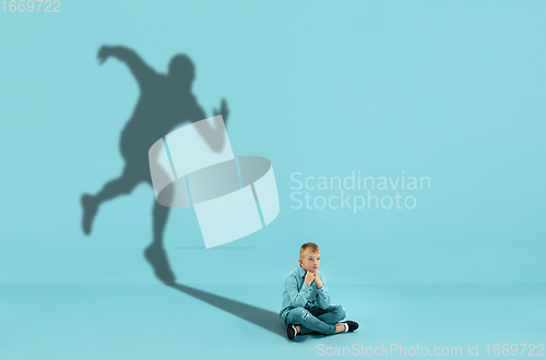 Image of Childhood and dream about big and famous future. Conceptual image with boy and shadow of fit male runner on blue background