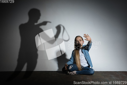 Image of Domestic violence, abusing. Scared little caucasian girl, victim sitting close to white wall with shadow of angry father\'s threatening on it.