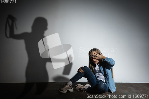 Image of Domestic violence, abusing. Scared little caucasian girl, victim sitting close to white wall with shadow of angry threatening mother with alcohol addiction.