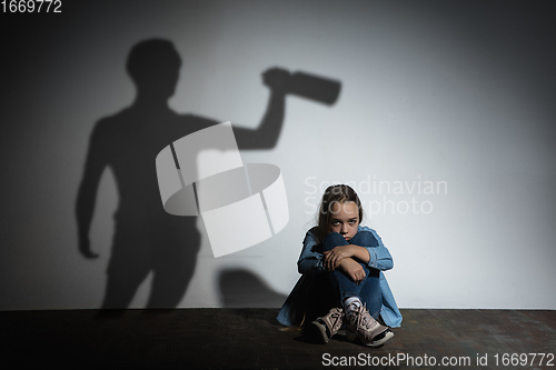 Image of Domestic violence, abusing. Scared little caucasian girl, victim sitting close to white wall with shadow of angry threatening father with alcohol addiction.