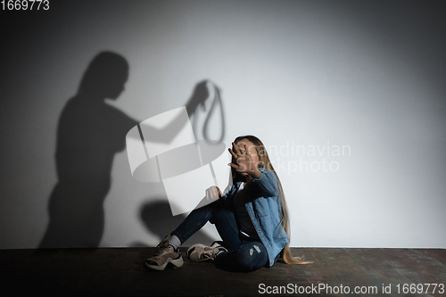 Image of Domestic violence, abusing. Scared little caucasian girl, victim sitting close to white wall with shadow of angry mother\'s threatening on it.