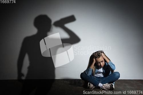 Image of Domestic violence, abusing. Scared little caucasian girl, victim sitting close to white wall with shadow of angry threatening father with alcohol addiction.