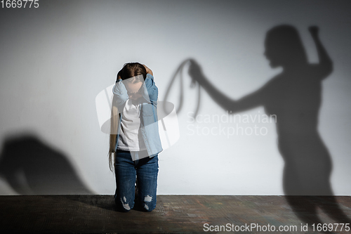 Image of Domestic violence, abusing. Scared little caucasian girl, victim sitting close to white wall with shadow of angry mother\'s threatening on it.