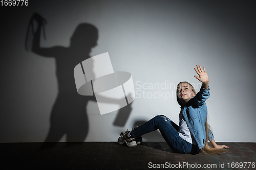 Image of Domestic violence, abusing. Scared little caucasian girl, victim sitting close to white wall with shadow of angry threatening mother with alcohol addiction.