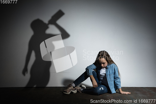 Image of Domestic violence, abusing. Scared little caucasian girl, victim sitting close to white wall with shadow of angry threatening mother with alcohol addiction.