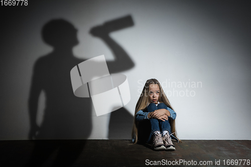 Image of Domestic violence, abusing. Scared little caucasian girl, victim sitting close to white wall with shadow of angry threatening father with alcohol addiction.