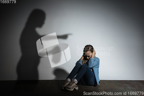 Image of Domestic violence, abusing. Scared little caucasian girl, victim sitting close to white wall with shadow of angry threatening mother with alcohol addiction.