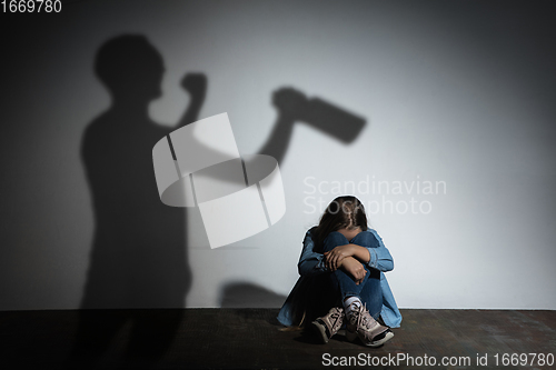 Image of Domestic violence, abusing. Scared little caucasian girl, victim sitting close to white wall with shadow of angry threatening father with alcohol addiction.