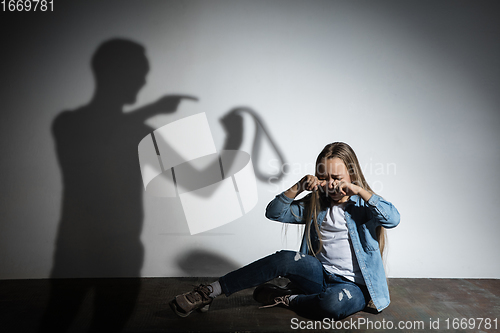 Image of Domestic violence, abusing. Scared little caucasian girl, victim sitting close to white wall with shadow of angry father\'s threatening on it.