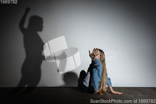 Image of Domestic violence, abusing. Scared little caucasian girl, victim sitting close to white wall with shadow of angry threatening mother with alcohol addiction.