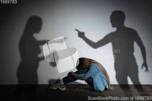 Image of Domestic violence, abusing. Scared little caucasian girl, victim sitting close to white wall with shadow of angry threatening parents with alcohol addiction.