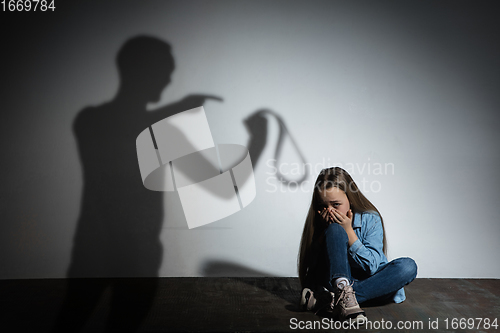 Image of Domestic violence, abusing. Scared little caucasian girl, victim sitting close to white wall with shadow of angry father\'s threatening on it.