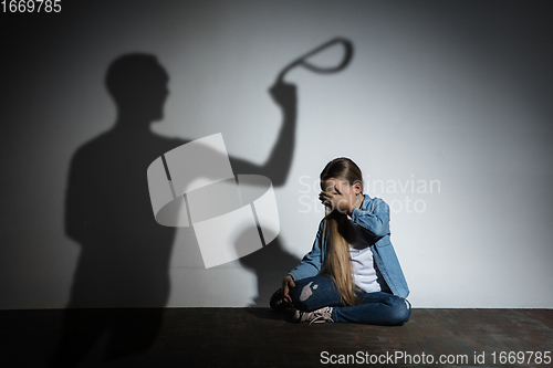 Image of Domestic violence, abusing. Scared little caucasian girl, victim sitting close to white wall with shadow of angry father\'s threatening on it.