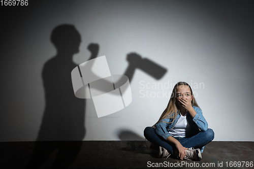 Image of Domestic violence, abusing. Scared little caucasian girl, victim sitting close to white wall with shadow of angry threatening father with alcohol addiction.