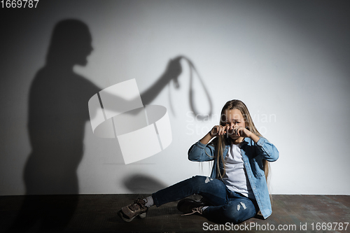 Image of Domestic violence, abusing. Scared little caucasian girl, victim sitting close to white wall with shadow of angry mother\'s threatening on it.