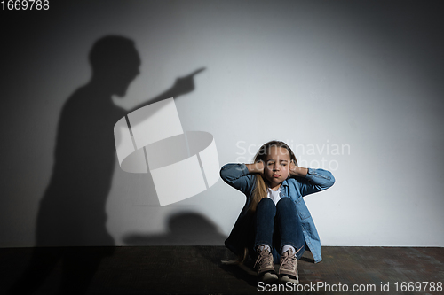 Image of Domestic violence, abusing. Scared little caucasian girl, victim sitting close to white wall with shadow of angry father\'s threatening on it.